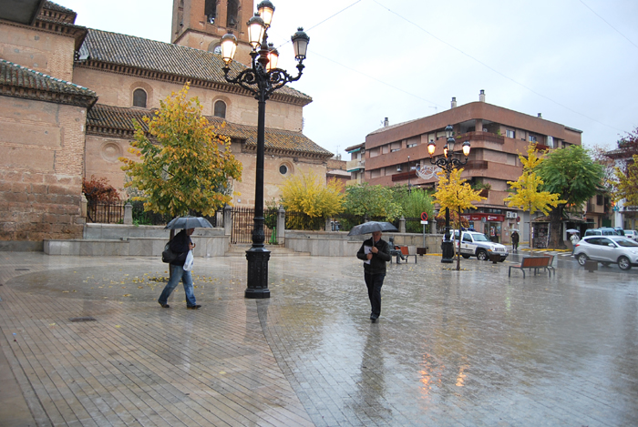 Dos viandantes se refugian con sus paraguas a su paso por la Plaza de España de Albolote 
